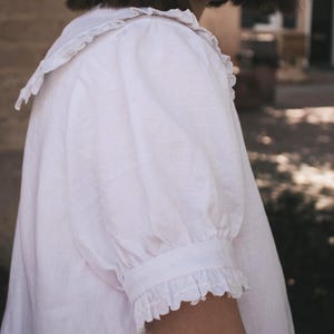 Puede incluir: Una blusa blanca que presenta un cuello grande con bordes con volantes, mangas cortas abullonadas y pu&ntilde;os con volantes a juego. La prenda est&aacute; confeccionada con un tejido texturizado y muestra un dise&ntilde;o sencillo con costuras visibles en las mangas.