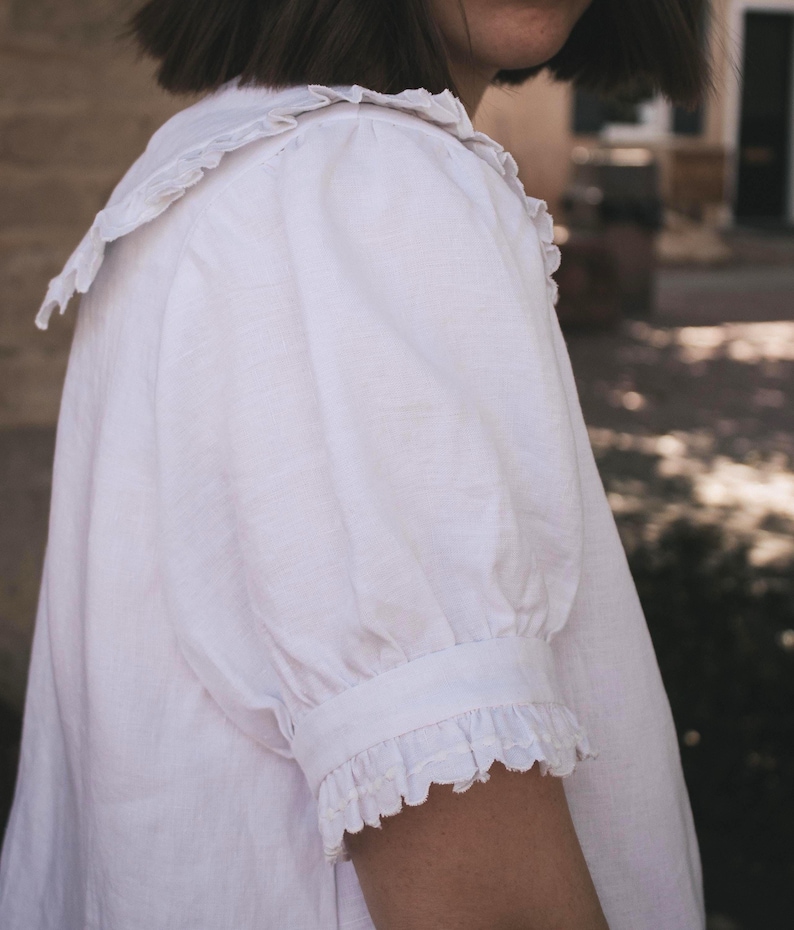 Puede incluir: Una blusa blanca que presenta un cuello grande con bordes con volantes, mangas cortas abullonadas y pu&ntilde;os con volantes a juego. La prenda est&aacute; confeccionada con un tejido texturizado y muestra un dise&ntilde;o sencillo con costuras visibles en las mangas.