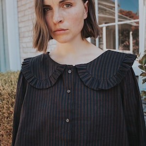 Puede incluir: Una camisa de manga larga con un patr&oacute;n de rayas verticales en marr&oacute;n oscuro y negro, un cuello ancho y redondeado con bordes con volantes y un cierre frontal de botones con tres botones oscuros visibles en el centro del pecho.