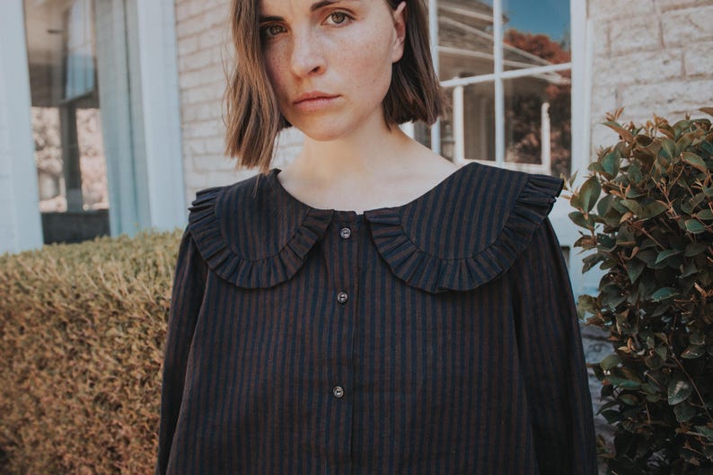 Puede incluir: Una camisa de manga larga con un patr&oacute;n de rayas verticales en marr&oacute;n oscuro y negro, un cuello ancho y redondeado con bordes con volantes y un cierre frontal de botones con tres botones oscuros visibles en el centro del pecho.
