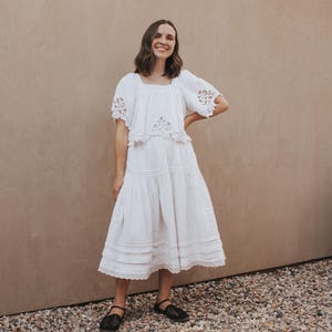 May include: A white dress with a square neckline and short sleeves with lace detailing. The dress has a tiered skirt and is paired with black flats. The background is a neutral tan color.