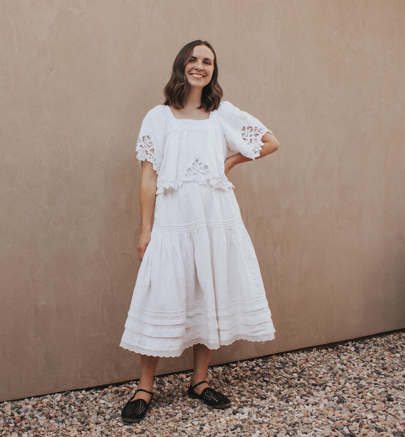 May include: A white dress with a square neckline and short sleeves with lace detailing. The dress has a tiered skirt and is paired with black flats. The background is a neutral tan color.