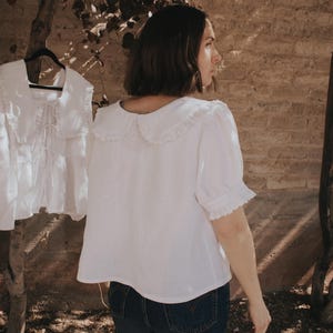 Puede incluir: Una blusa blanca de manga corta con un cuello grande y redondeado con bordes de volantes y pu&ntilde;os a juego tambi&eacute;n con volantes. La blusa tiene un corte holgado y recto, combinada con vaqueros azul oscuro. Una segunda blusa id&eacute;ntica cuelga cerca.