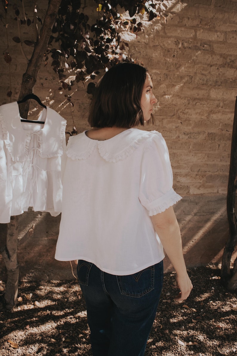 Puede incluir: Una blusa blanca de manga corta con un cuello grande y redondeado con bordes de volantes y pu&ntilde;os a juego tambi&eacute;n con volantes. La blusa tiene un corte holgado y recto, combinada con vaqueros azul oscuro. Una segunda blusa id&eacute;ntica cuelga cerca.