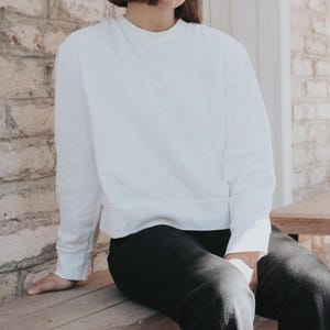 May include: A white long-sleeved shirt with a crew neck and a relaxed fit. The shirt is tucked into black pants.