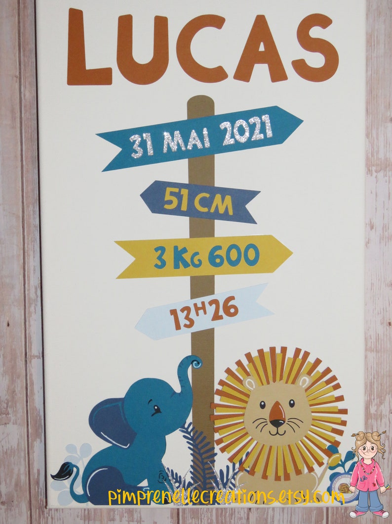 K&ouml;nnte beinhalten: Ein wei&szlig;es Leinwand-Schild mit dem Namen "Lucas" in orangefarbenen Buchstaben. Das Schild hat blaue, gelbe und hellblaue Pfeile mit dem Text "31 Mai 2021", "51 cm", "3 kg 600" und "13h26". Das Schild zeigt auch einen blauen Elefanten und einen gelben L&ouml;wen.