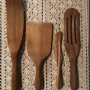 May include: Four wooden kitchen utensils, including a spatula, a slotted spatula, a butter knife, and a large spoon, are arranged vertically on a beige crocheted surface.