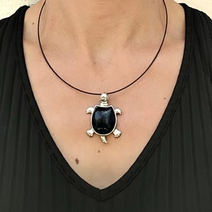 Peut inclure: Un pendentif tortue de couleur argentée doté d'une incrustation ovale noire lisse et polie pour la carapace. Le pendentif est suspendu à un collier fin et sombre en forme de cercle qui repose autour du cou, créant un bijou simple et épuré.