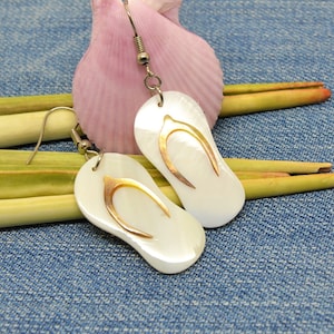 Op de afbeelding: Een paar witte schelp-oorbellen in de vorm van slippers met gouden accenten. De oorbellen hangen aan zilveren haakjes.