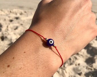 Puede incluir: Pulsera de hilo rojo con una cuenta azul de ojo turco y detalles dorados. La pulsera se lleva en la muñeca, con una playa y el océano de fondo.