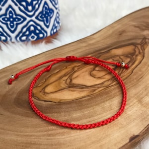 May include: A bright red woven bracelet with adjustable sliding knots and small gold-colored beads. The bracelet is displayed on a wooden surface with a natural grain pattern, with a blue and white patterned ceramic object in the background.
