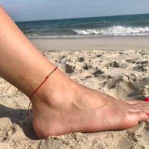 Może przedstawiać: Czerwona bransoletka na kostkę z małymi złotymi koralikami. Bransoletka jest noszona na stopie na piaszczystej plaży z oceanem w tle. Paznokcie u stóp osoby są pomalowane na czerwono.