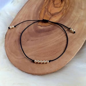 May include: A black cord bracelet with gold-colored beads. The bracelet features a sliding knot closure for adjustable sizing. The beads are evenly spaced along the cord, creating a minimalist design.