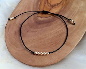 May include: A black cord bracelet with gold-colored beads. The bracelet features a sliding knot closure for adjustable sizing. The beads are evenly spaced along the cord, creating a minimalist design.