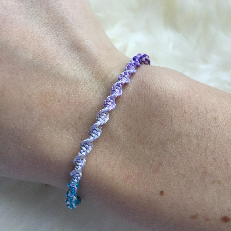 May include: A handmade bracelet with a spiral design. The bracelet features a gradient of colors, transitioning from blue to purple. The bracelet is made of woven threads, creating a textured appearance. The bracelet is designed to be worn on the wrist.