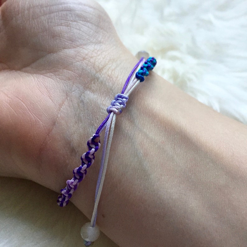 May include: A handmade bracelet featuring a macrame design with purple, white, and teal threads. The bracelet is knotted and has a sliding knot closure. A small, round, white bead is at the end of the bracelet.