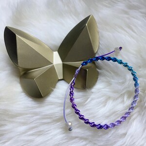 May include: A handmade macrame bracelet with a gradient of purple, blue, and turquoise colors. The bracelet is next to a gold-colored butterfly-shaped decorative object. The background is a soft, white, fluffy surface.