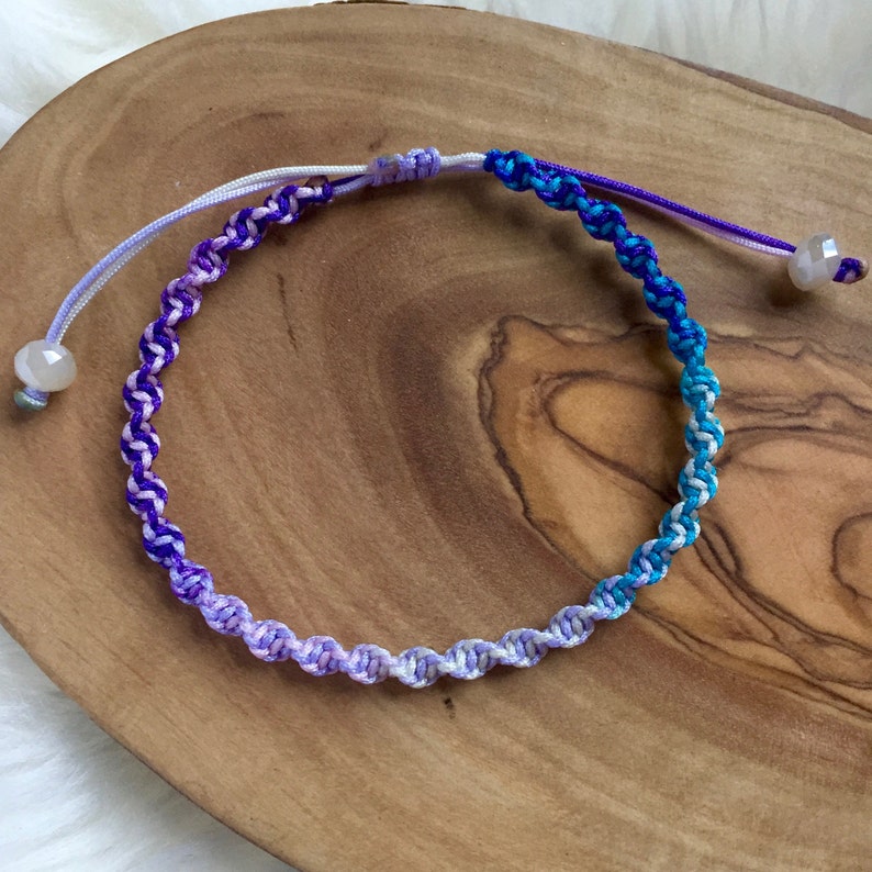 May include: A handmade macrame bracelet with a gradient of colors, transitioning from light purple to deep blue. The adjustable bracelet features a sliding knot closure and a small, iridescent bead at each end.
