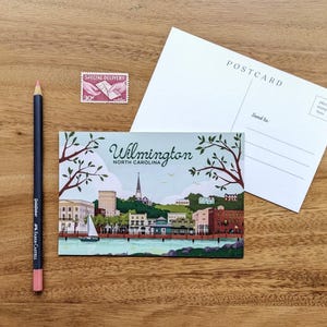 May include: A postcard featuring a colorful illustration of Wilmington, North Carolina, with a sailboat on the water. A pink pencil, a postage stamp, and a blank postcard are also on a wooden surface.