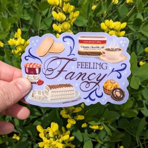 May include: A sticker with a purple background and the text "Feeling Fancy" in a decorative font. The sticker features illustrations of a cup of coffee, a chocolate bar, a jar of jam, a pastry, and a chocolate candy.