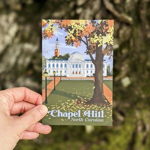 A postcard with a retro-inspired design of the UNC campus is held in front of a tree trunk. The design shows Polk Place quad in the fall.
