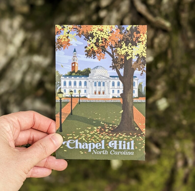 A postcard with a retro-inspired design of the UNC campus is held in front of a tree trunk. The design shows Polk Place quad in the fall.