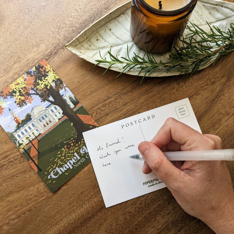 A hand is shown writing a message on a postcard with a retro-inspired design of the UNC campus. Chapel Hill, North Carolina is printed at the bottom in a blue retro font.