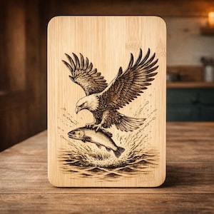 May include: A rectangular wooden cutting board with rounded corners. The board features a detailed wood-burned image of a bald eagle with outstretched wings perched atop a fish, with water splashing around them. The wood has a natural, light brown color.