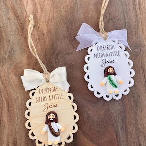 May include: Two decorative ornaments with scalloped edges and the text "EVERYBODY NEEDS A LITTLE Jesus". One is wood, the other white. Each features a small Jesus figure with a brown robe and a white tunic, and a ribbon for hanging.