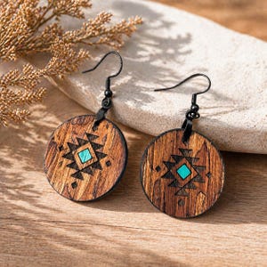 May include: Wooden earrings with a circular design. The earrings feature a dark wood grain pattern with a black geometric design and a turquoise square in the center. The earrings have black metal hooks and leather accents.