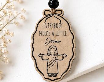 May include: An oval-shaped wooden hanging ornament with a scalloped edge, featuring a black printed illustration of Jesus with arms outstretched. The text reads, "EVERYBODY NEEDS A LITTLE Jesus" and "Jesus loves you" on the robe, suspended by a brown cord with a black bead.
