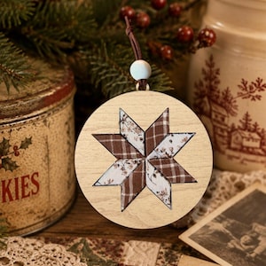 May include: A circular wooden ornament featuring a central eight-pointed star design. The star is composed of alternating fabric segments in brown plaid and a white floral pattern. A small white bead and a brown leather cord are attached at the top.