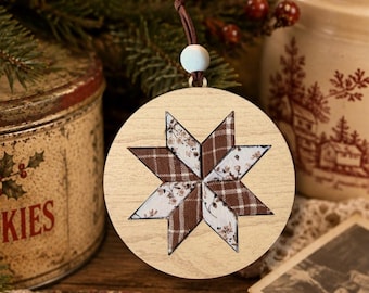May include: A circular wooden ornament featuring a central eight-pointed star design. The star is composed of alternating fabric segments in brown plaid and a white floral pattern. A small white bead and a brown leather cord are attached at the top.