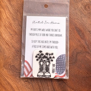 May include: A clear plastic package containing a white card with text and a small, white, rectangular pocket hug. The pocket hug features a black and white illustration of combat boots with flowers. The card has patriotic imagery.