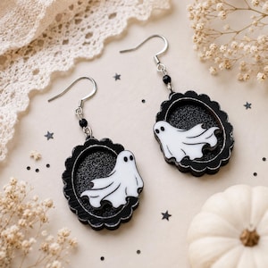 May include: A pair of dangle earrings featuring white ghost figures set against a black glittery background within scalloped black oval frames. The charms hang from silver-toned metal hooks accented with small black beads.