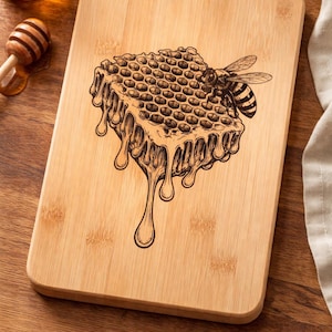 Könnte beinhalten: Rechteckiges Schneidebrett aus Bambus mit einem gravierten Design einer Honigwabe mit tropfendem Honig und einer Biene. Das Brett ist hellbraun und hat abgerundete Ecken. Ein Honigglas und ein Honiglöffel sind sichtbar.