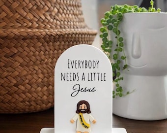 May include: A white decorative sign with the text "EVERYBODY NEEDS A LITTLE Jesus" and a small Jesus figurine. The figurine has brown hair and a yellow sash that says "Jesus loves you". The sign is on a white base.