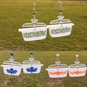 Puede incluir: Pendientes con miniaturas de fuentes blancas para horno con tapas transparentes. Los pendientes muestran varios diseños florales en verde, azul y naranja. Cada pendiente está suspendido de un gancho plateado con cuentas decorativas.