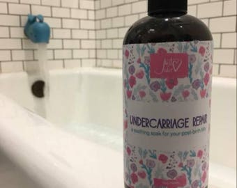 May include: A dark brown plastic bottle with a black cap featuring a white label with a pink and blue floral pattern. The label text reads Just Julie, UNDERCARRIAGE REPAIR, and a soothing soak for your post-birth bits in dark purple and blue.