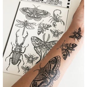 May include: A sheet of black and white temporary tattoos from J'IRIS STUDIO TEMPORARY TATTOO COLLECTIONS featuring detailed illustrations of insects including moths, a beetle, a dragonfly, a scorpion, and bees, with three applied to skin.