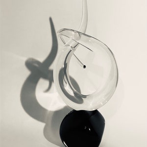 Op de afbeelding: Een helder glazen sculptuur met een wervelend, abstract ontwerp. Het beeld is gemonteerd op een zwarte, ronde basis.