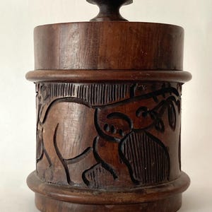 Könnte beinhalten: Ein dunkelbrauner Holzbehälter mit Deckel. Der zylindrische Behälter weist ein geschnitztes Relief eines Elefanten und Blättern auf. Der Deckel hat einen kleinen, abgerundeten Knopf. Wahrscheinlich zur Aufbewahrung verwendet.