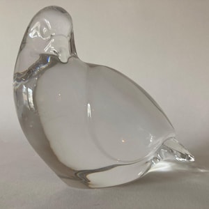 Op de afbeelding: Een helder glazen duifbeeldje met een witte, ondoorzichtige body. De vogel is gebeeldhouwd met een gladde, afgeronde vorm, met een gedetailleerde kop en staart. Het glas reflecteert licht, waardoor highlights en schaduwen ontstaan, en is een decoratief item.