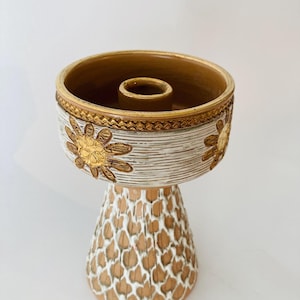 Op de afbeelding: Een decoratieve kandelaar met een voetstuk. De bovenkant heeft de vorm van een kom met een gouden rand en een centraal gat voor een kaars. De buitenkant is wit met gouden bloemaccenten. De basis is bruin met een wit patroon.