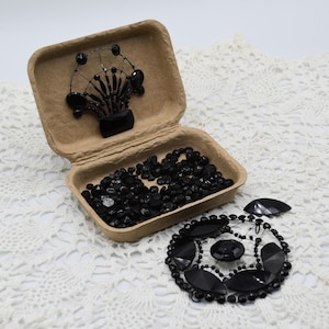 Peut inclure: Un récipient en carton ouvert rempli de perles noires et d'une composition florale perlée noire. Un ornement perlé noir et des perles en vrac sont également visibles sur une surface en dentelle blanche.