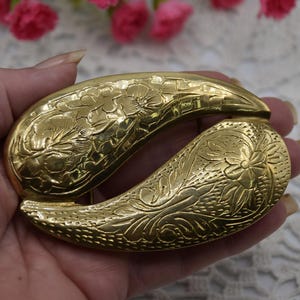 May include: A gold-coloured, vintage belt buckle with an ornate floral and leaf design. The buckle is a curved, double-ended shape, with detailed embossed patterns. The metal has a polished finish, suggesting a classic or antique style.
