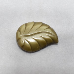 May include: A green leaf-shaped button with a raised design. The button is made of plastic and has a glossy finish.