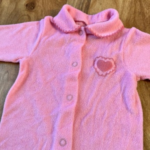 May include: A pink baby onesie with a heart embroidered on the front. The onesie has a button closure and a ruffled collar.