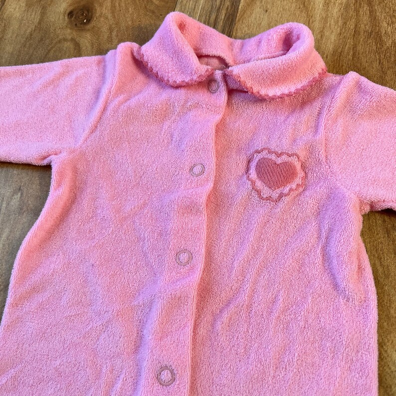 May include: A pink baby onesie with a heart embroidered on the front. The onesie has a button closure and a ruffled collar.