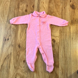 May include: A pink baby onesie with a heart-shaped design on the chest. The onesie has a button closure down the front and a collar.